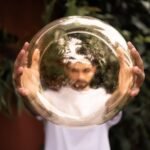A man holds a reflective golden sphere, creating a distorted image of his face outdoors.