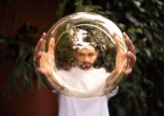 A man holds a reflective golden sphere, creating a distorted image of his face outdoors.