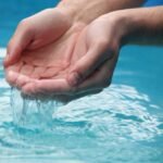 Close-up of hands scooping clear blue water from a reflective pool outdoors.
