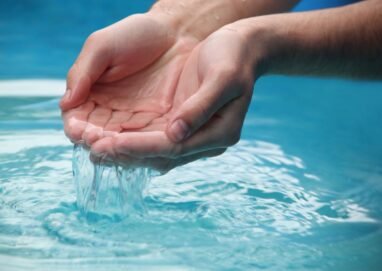 Close-up of hands scooping clear blue water from a reflective pool outdoors.