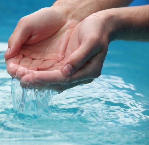 Close-up of hands scooping clear blue water from a reflective pool outdoors.