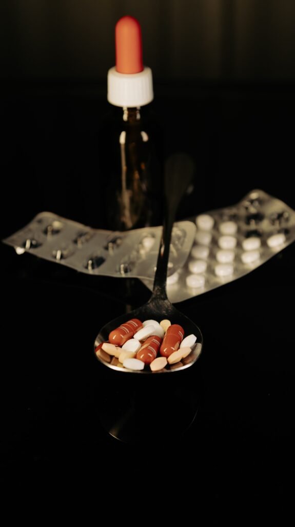 Close-up of assorted pills on a spoon with blister packs and a medicine dropper in the background.
