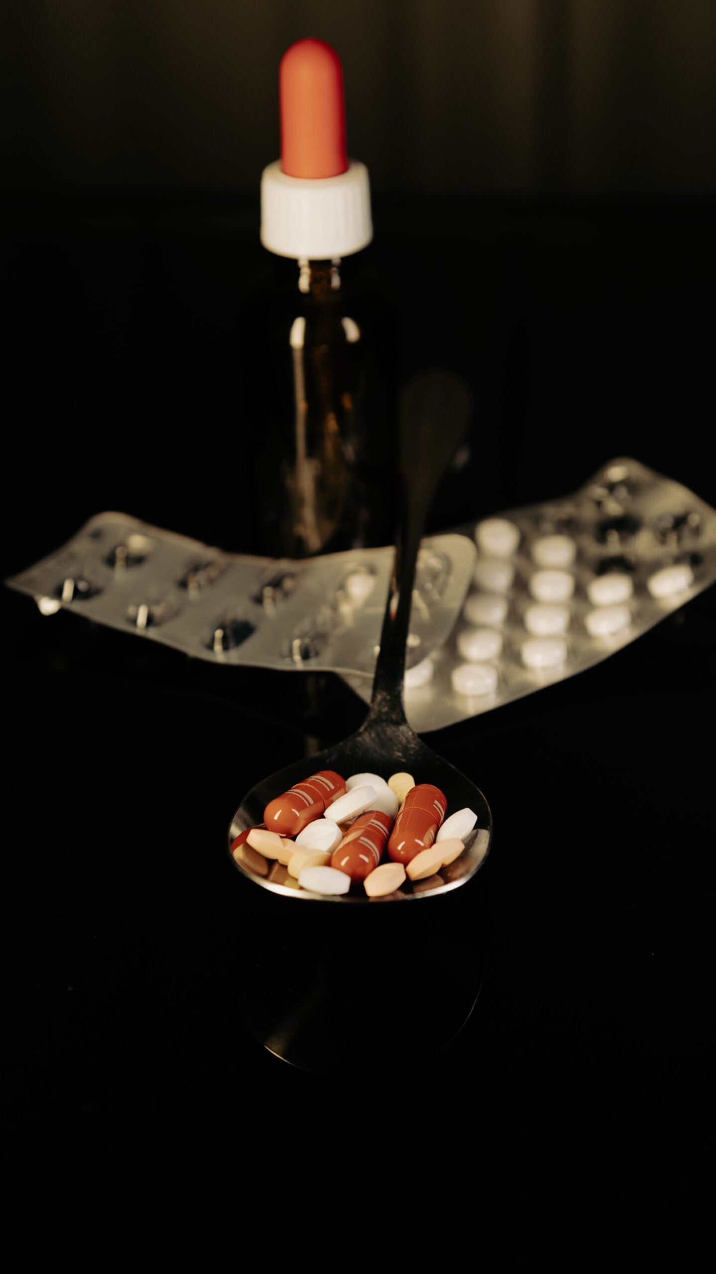Close-up of assorted pills on a spoon with blister packs and a medicine dropper in the background.