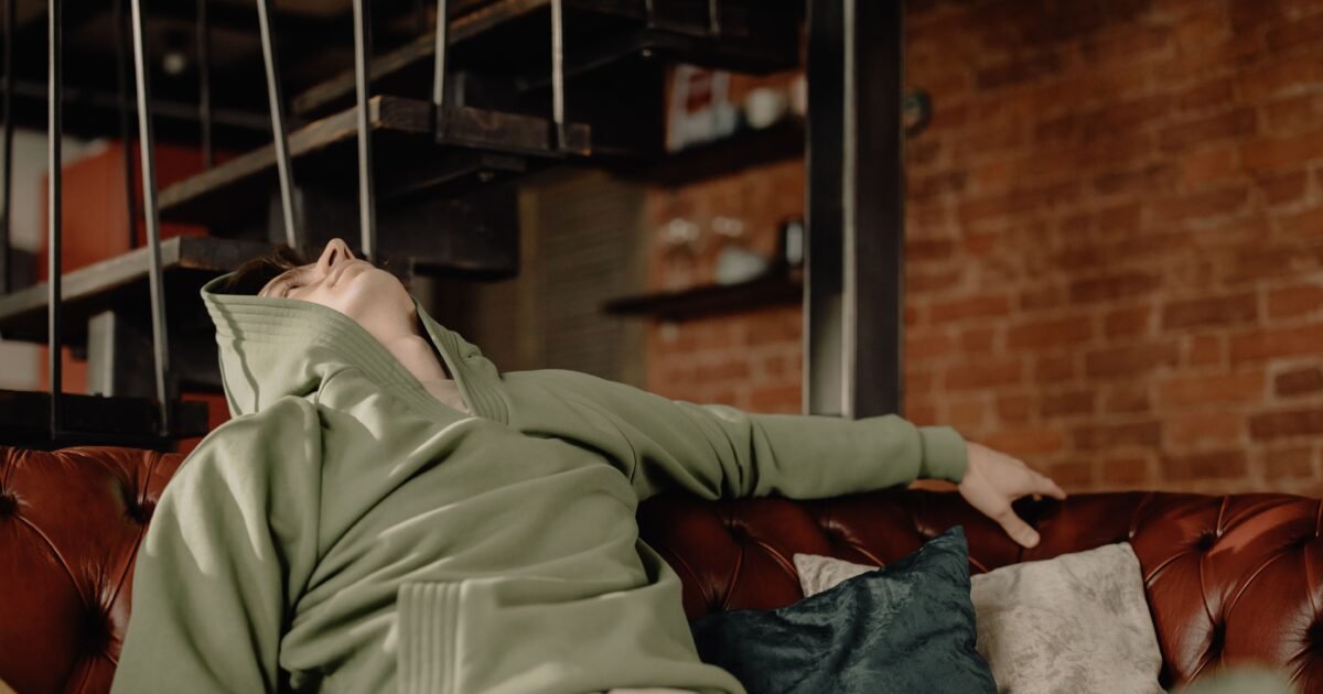 A young man in a green hoodie relaxes on a leather sofa at home, embodying a cozy and laid-back atmosphere.