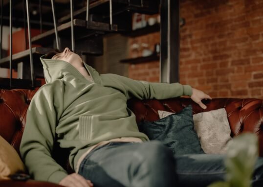 A young man in a green hoodie relaxes on a leather sofa at home, embodying a cozy and laid-back atmosphere.