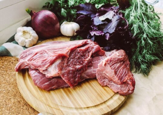 Close-up of raw beef on a wooden board with fresh herbs and vegetables.