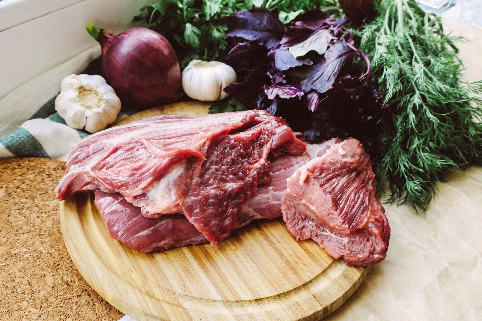 Close-up of raw beef on a wooden board with fresh herbs and vegetables.