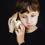A young boy with brown hair listens to a large seashell against a black background.