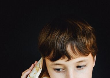 A young boy with brown hair listens to a large seashell against a black background.