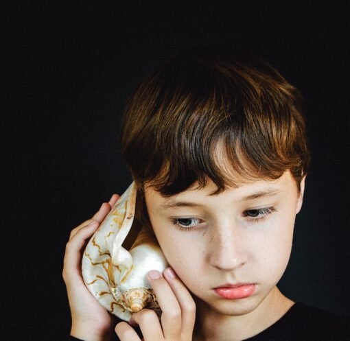 A young boy with brown hair listens to a large seashell against a black background.