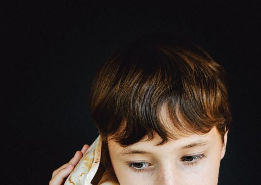 A young boy with brown hair listens to a large seashell against a black background.