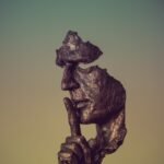 Bronze sculpture of a face missing parts and a finger to its lips, suggesting silence and mystery.