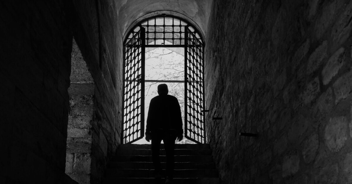 A solitary silhouette in an ancient stone corridor with a gated archway.