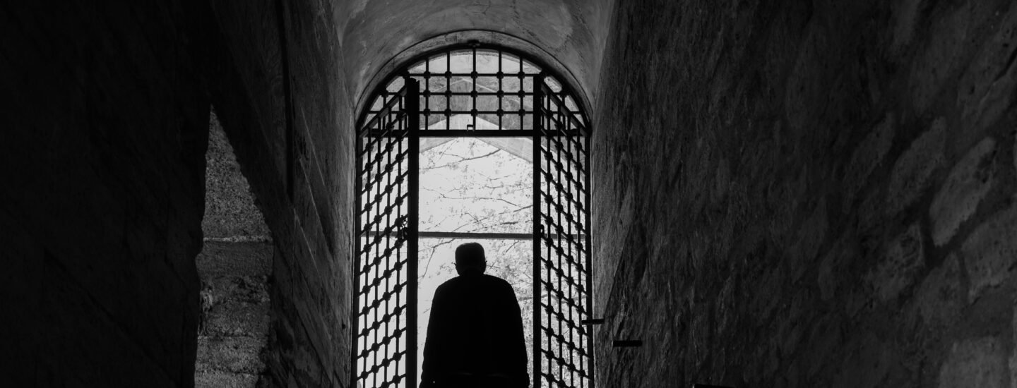 A solitary silhouette in an ancient stone corridor with a gated archway.