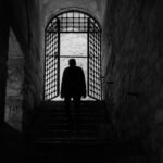 A solitary silhouette in an ancient stone corridor with a gated archway.