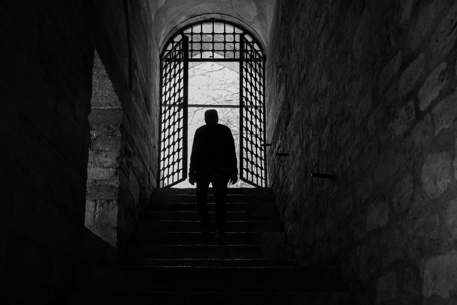 A solitary silhouette in an ancient stone corridor with a gated archway.