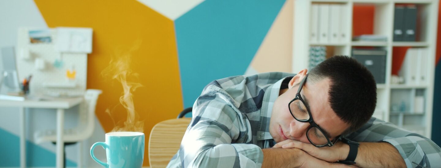 Young man sleeping on a laptop at his desk with a hot beverage nearby.