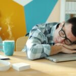 Young man sleeping on a laptop at his desk with a hot beverage nearby.