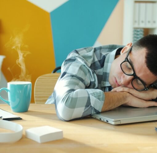 Young man sleeping on a laptop at his desk with a hot beverage nearby.