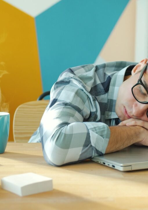 Young man sleeping on a laptop at his desk with a hot beverage nearby.
