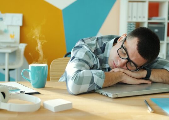 Young man sleeping on a laptop at his desk with a hot beverage nearby.