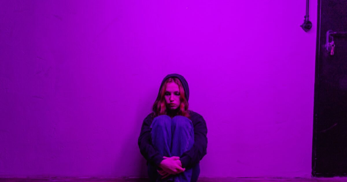 A woman in a hoodie sits alone on the floor, illuminated by purple light indoors.