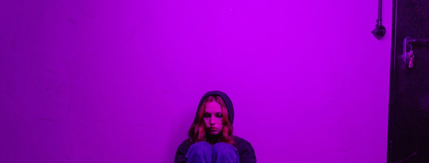 A woman in a hoodie sits alone on the floor, illuminated by purple light indoors.