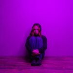 A woman in a hoodie sits alone on the floor, illuminated by purple light indoors.