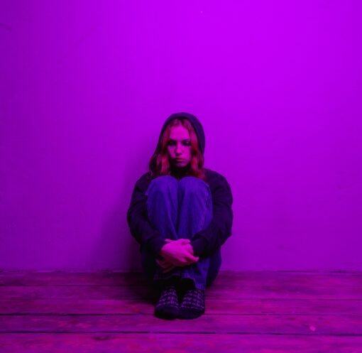 A woman in a hoodie sits alone on the floor, illuminated by purple light indoors.