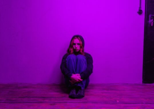 A woman in a hoodie sits alone on the floor, illuminated by purple light indoors.