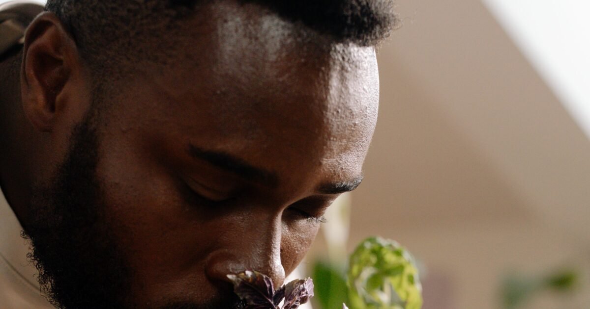 A close-up of a bearded man with eyes closed, enjoying the scent of a potted plant indoors.